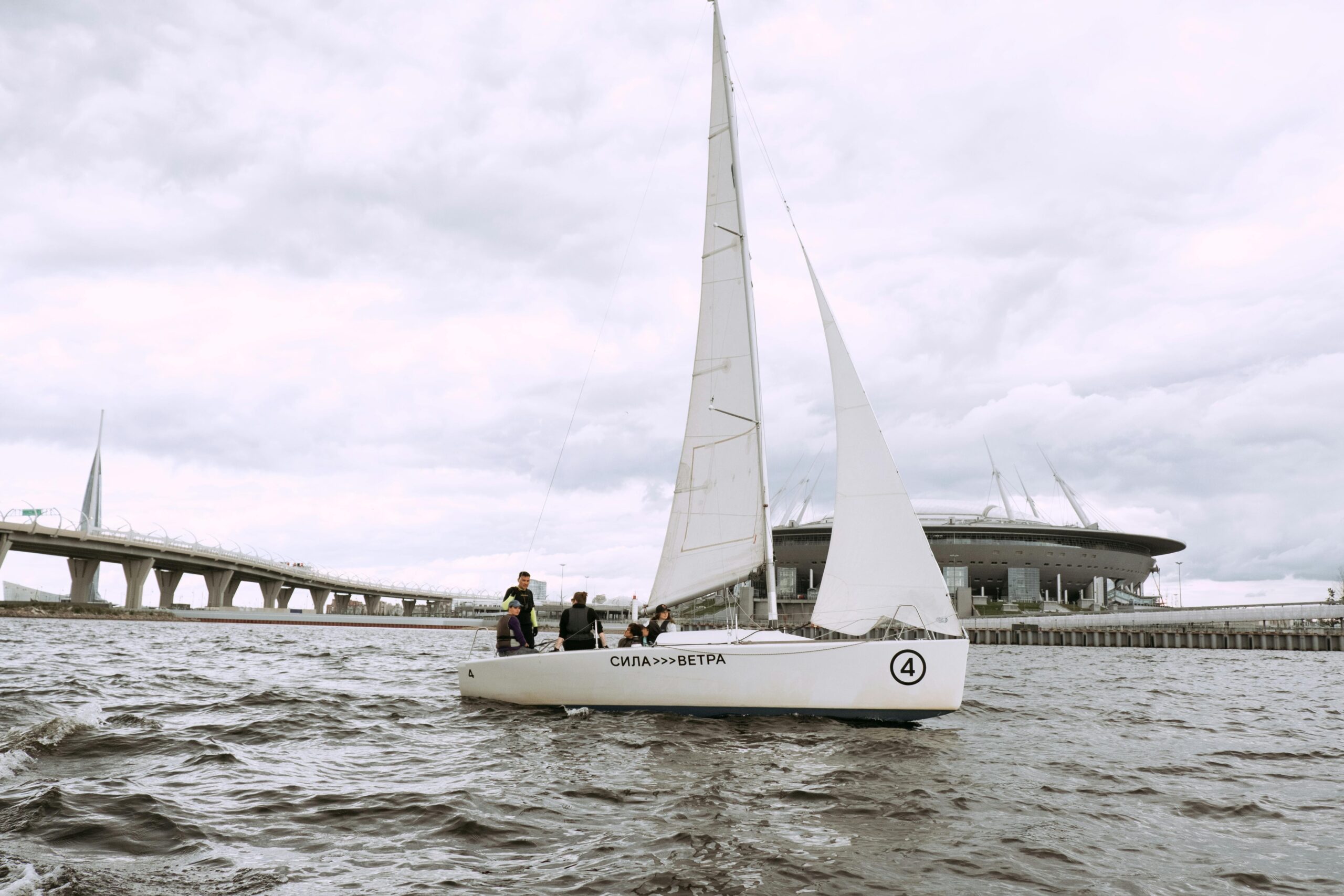 A yacht sailing near a modern bridge and stadium under cloudy skies.