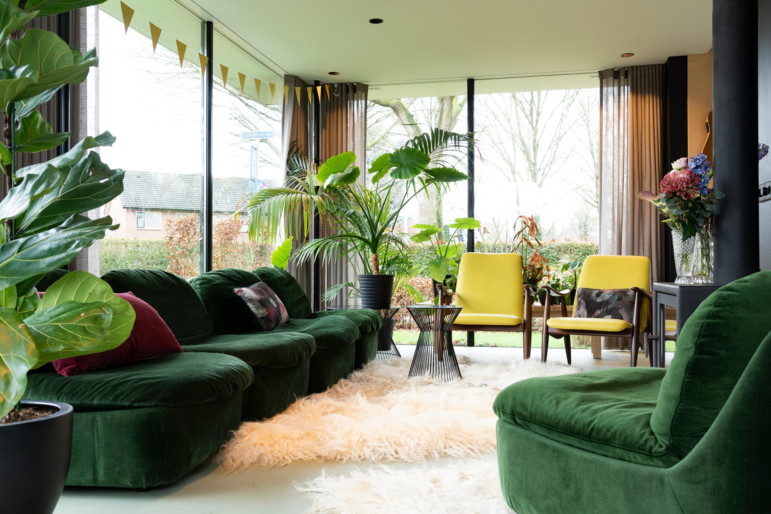 A modern living room featuring a green sofa, yellow chairs, and lush indoor plants, bathed in natural light.