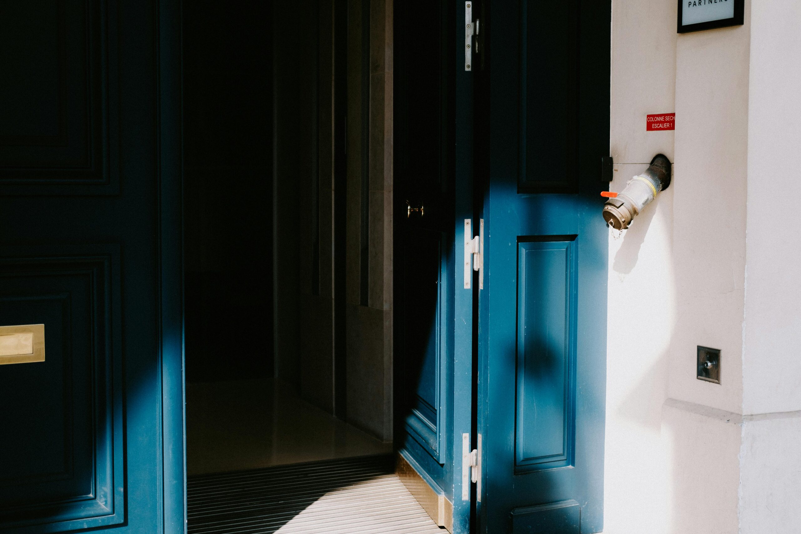 A sunlit blue door slightly ajar in a residential building entrance, casting shadows.