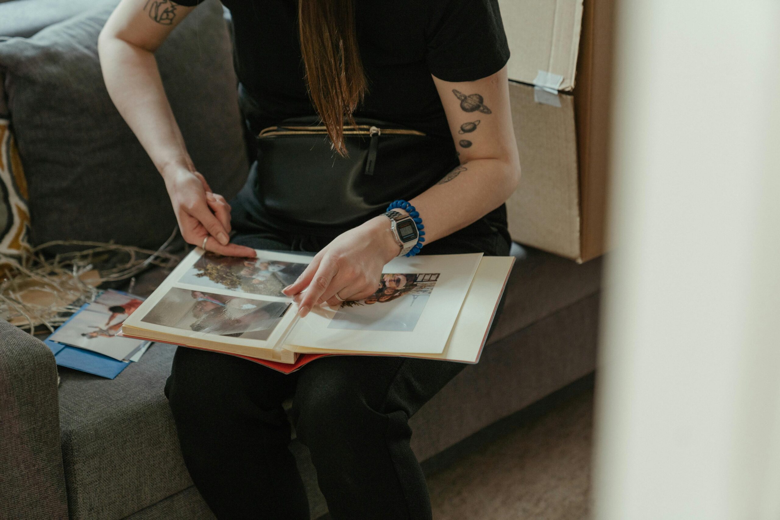 A person with tattoos sits indoors examining a photo album on a sofa.